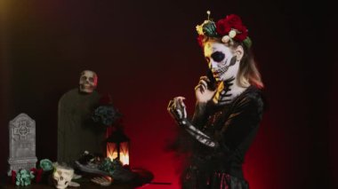 Santa Muerte modeli akıllı telefondan konuşuyor, cadılar bayramı makyajı yapıyor ve stüdyoda çiçek tacını takıyor. La Cavalera Catrina, Dios de los Muertos Meksika tatili hakkında konuşmak için akıllı telefonu kullanıyor..