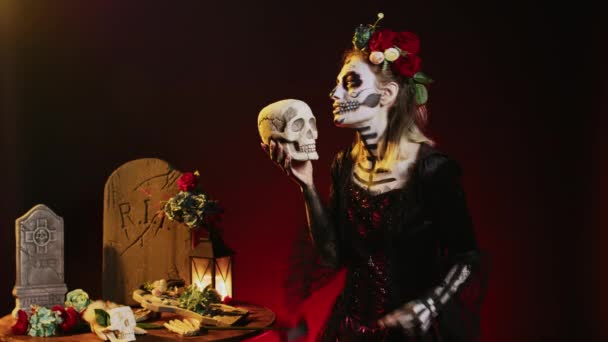 スタジオで神聖な頭蓋骨を持っている不気味なグラマラスな女性は メキシコのハロウィーンの日を祝うために怖いと恐怖を演じています 体の芸術と祭りの衣装を身に着けている薄汚い女神 死の女性のように見えます — ストック動画