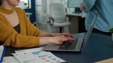 Portrait of woman typing using laptop keyboard and smiling at camera sitting at desk in busy startup office. Casual business employee working relaxed with papers with charts and statstics.
