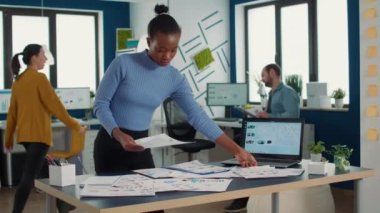 African american startup employee looking at business papers with data charts comparing two sheets with statistics on laptop screen. Woman standing in finance department analyzing profit indicators.
