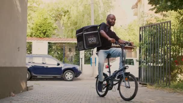 男性配達宅配便顧客にファーストフード注文パッケージを与えるために熱バックパック付き自転車に乗る 玄関前でテイクアウト食事やレストランデリバリーサービスを提供します — ストック動画