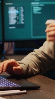 Dikey video: Bilişim dolandırıcılığı suçlusu siber güvenlik sistemini kırıp birden fazla monitörden veri çalıyor. Kapüşonlu tehlikeli bir hacker kahve içiyor ve bilgisayar sunucusuna kötü yazılım yüklüyor.