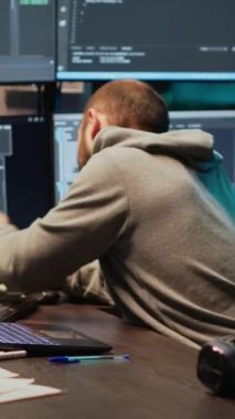 Dikey video: Yazılım geliştiricisi geceleri sabit hattan konuşuyor, ofisteki telefonlara cevap veriyor. Çevrimiçi bulut hesaplaması üzerinde çalışmak ve sohbet etmek için telefonla telefon kullanan erkek programcı, yapay