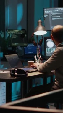 Dikey video: Genç yetişkin kodlayıcı gece telefonda konuşuyor, ofisteki telefona cevap veriyor. Çevrimiçi bulut hesaplaması üzerinde çalışmak ve sohbet etmek için telefonla telefon kullanan erkek programcı, yapay