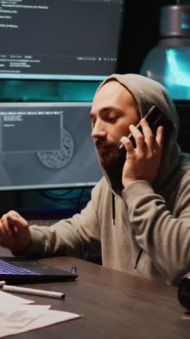 Dikey video: Bilgi teknolojileri geliştirme ofisinde telefonla konuşan genç programcı, uzaktan konuşma için akıllı telefonu kullanıyor. Html kodu üzerinde çalışan erkek mühendis geliştiriyor, cep telefonu tutuyor. El bilgisayarı