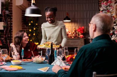 Aile Noel kutlaması, şenlikli yemek masası geleneksel tavuğu elinde tutan gülen kadın. Mutlu insanlar kostüm yiyor, kış tatili ev partisi veriyor.