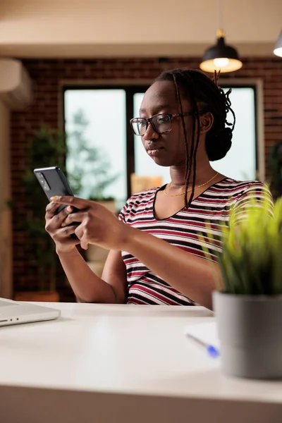 若いアフリカ系アメリカ人女性がスマートフォンでメッセージを入力し 職場のソーシャルメディアでチャットします 携帯電話でインターネットをサーフィンするフリーランスは 自宅のオフィスの机に座って — ストック写真