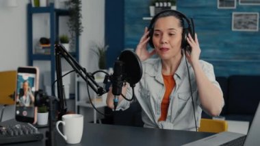 Happy influencer starting to record daily vlog while putting on headphones in home studio. Beautiful social media star recording video for audience while having professional audio equipment.