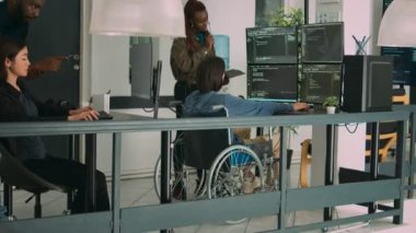 Fiziksel engelli Asyalı kodlayıcı terminalin penceresinde güvenlik duvarı oluşturmak için Afrikalı Amerikalı geliştiriciyle çalışıyor. Tekerlekli sandalye programlama html kod mühendisi.