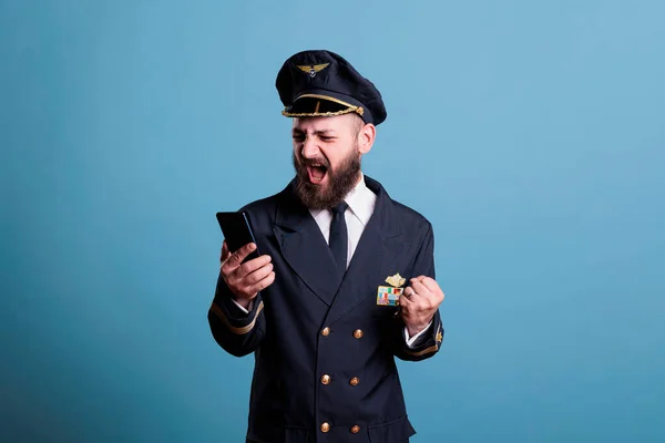 勝者のジェスチャーで近代的な携帯電話を保持制服を着た幸せな笑顔の飛行機のパイロットは 拳を食いしばった 携帯電話の画面を見て陽気な表情のパイロット 良いニュースを読む — ストック写真