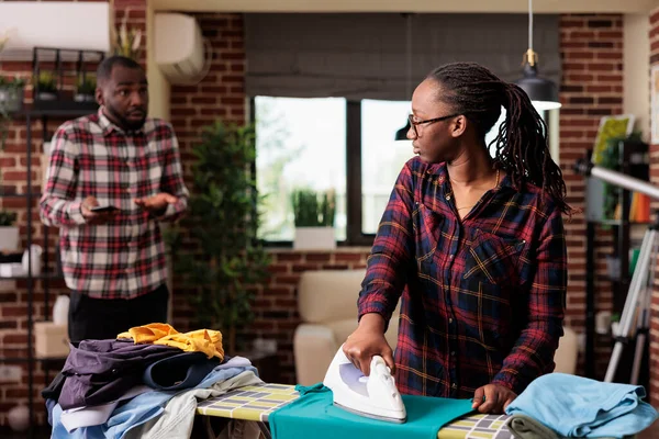気が散る夫は携帯電話を使いながら一人で家事をする人妻 アフリカ系アメリカ人のカップルが同じ責任について話し合い 不満を抱く女性が家事を手伝う — ストック写真