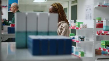 Afro-Amerikalı bir kadın eczane raflarındaki ilaç paketlerini kontrol ediyor, eczanedeki kutularda sağlık ürünleri ve vitaminler alıyor. Hap ve uyuşturucuya bakan kişi.