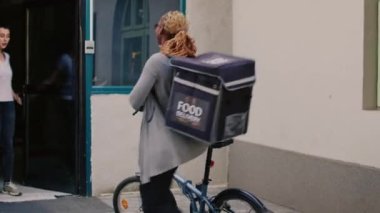 Yemek teslimatı kuryesi kadınlara kese kağıdında yemek veriyor, bisikletle restoran yemeği dağıtıyor. Bina girişinde paket servis etmek için bisiklete binmek ve sırt çantası taşımak..