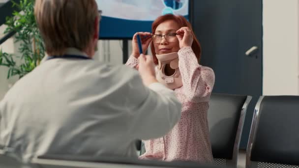 医師は 病院のロビーでアジアの女性と協議を行うと 子宮頸椎を着て 損傷後の脳損傷を確認するために視力検査を行うフォームブレースを有する患者 手持ち撮影 — ストック動画