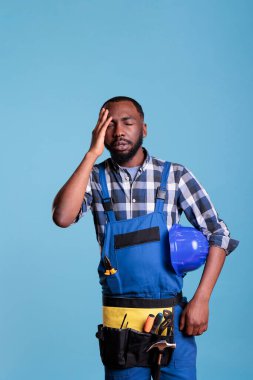 Üniformalı stresli müteahhit ve yorgun kasketli, başını ovuşturan yorgun ve başı ağrıyor. Hüsrana uğramış inşaat işçisi, çok çalıştığı ve mavi arka planda dinlenemediği için acı çekiyor..