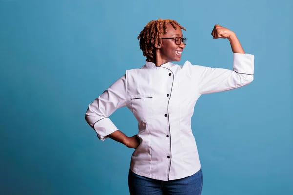 厨房女工作人员穿着厨师夹克 面带微笑地看着蓝色背景下的手臂肌肉 专业的厨房妇女在演播室拍摄时 以授权和力量的姿态弯曲着胳膊 — 图库照片