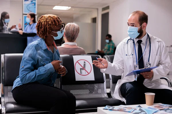 General medical practitioner explaining condition, illness to young african american female patient in hospital patient room. Physician attending sick woman, wearing cover masks for disease prevention