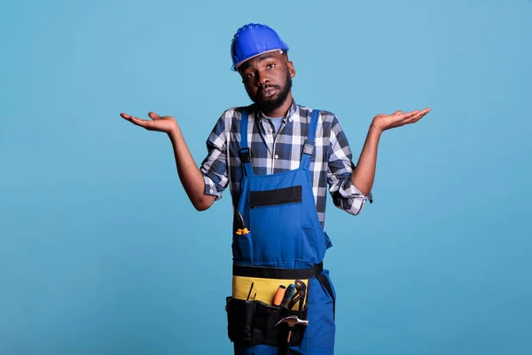 Uncertain and clueless builder making the I do not know gesture on camera, feeling doubtful and confused. Unsure man not getting the idea in studio shot against blue background.