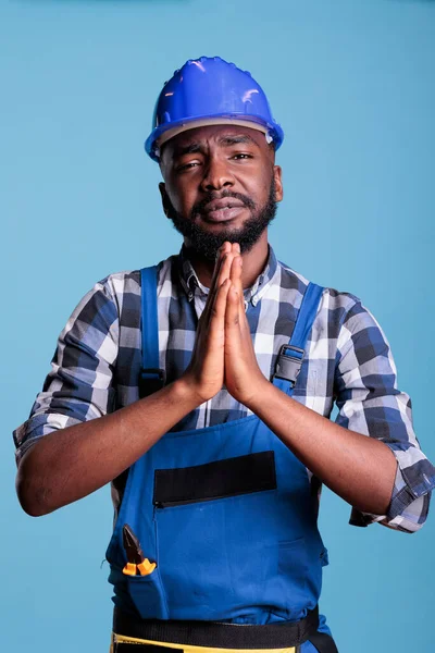 African american builder doing prayer hands on camera, begging and asking for good luck and fortune. Construction worker waiting for wish or request and imploring for desire, studio shot.
