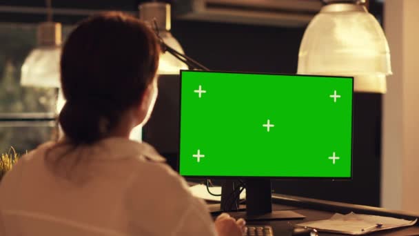 コンピュータ上の緑の画面のテンプレートで作業する女性は 空白のモックアップの背景を見て スタートアップ企業のオフィスの日没時に画面上の隔離されたクロマキーディスプレイと画面を分析 — ストック動画