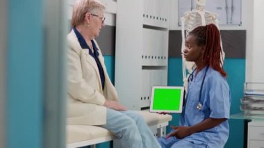 Nurse and senior patient looking at greenscreen on digital tablet in medical cabinet. Health specialist holding device with blank mockup template and isolated chromakey copyspace.