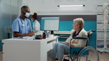 Male nurse consulting elderly woman with physical disability, doing checkup examination with senior patient during covid 19 pandemic. Talking to person in wheelchair about healthcare treatment.