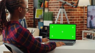 Laptop 'ta yeşil ekran görüntüsüne bakan kadın çalışan, kablosuz bilgisayarda boş model şablonu kullanıyor. Ana masadaki krom tuşlu kopya alanı arka planında çalışan kadın.
