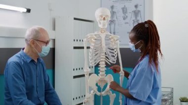 Yaşlı bir adamla insan iskeleti incelemesi yapan kadın hemşire, kabinedeki randevudaki osteopati teşhisini açıklıyor. Hastayla birlikte ortopedik omurilik anatomisine bakan tıbbi asistan..