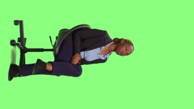 Dikey video: Ofis çalışanı sandalyede oturuyor ve stüdyoda bekliyor. Takım elbiseli genç iş kadını bekleyip hazırlanmak için yeşil perde arka planında bekliyor..