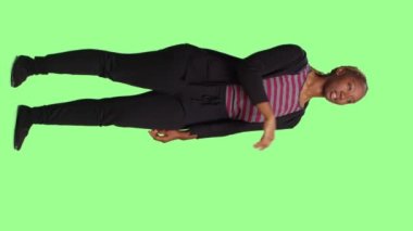 Dikey video: Holografik görüntü ve yapay zeka kullanarak yeşil ekran arkaplanından holograma bakan genç yetişkinler. Tam vücut üzerinde duran kadın modeli izole edilmiş yeşil ekran arkaplanı.