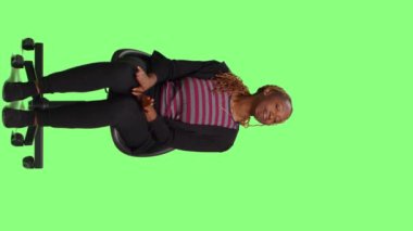 Dikey video: Stüdyoda sandalyede oturan kadın model tüm vücudu yeşil ekran arka planda, stüdyoda bekliyor ve hazırlanıyor. Sabırsız kadın taburede oturup bekliyor, yeşil ekran.