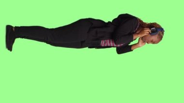 Dikey video: Genç mutlu yetişkin kulaklık kullanarak müzik dinliyor, tüm vücudu yeşil ekranda duruyor. MP3 radyo şarkıları ve ses kablosuz kulaklıklarıyla eğleniyorum..