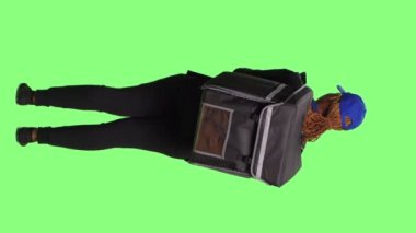 Dikey video: Kurye termal sırt çantasıyla yemek dağıtıyor ve akıllı telefondan interneti tarıyor, yeşil ekran arka planda duruyor. Müşterilere fast food siparişi vermek için internet sitesini kullanmak.