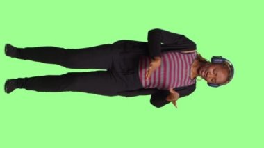 Dikey video: Kablosuz kulaklıkla müzik dinleyen güler yüzlü kendine güvenen bir kadın, tüm vücudu yeşil ekranda duruyor. MP3 radyo şarkıları ve ses kulaklıklarıyla eğlenmek, oyunculuk.