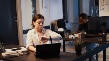 Ofis çalışanı geceleri dizüstü bilgisayarda çalışıp kol saatini kontrol ediyor, saate bakıyor ve mesai sonrası işte oluyor. Web belgeleri planlayan ve e-posta gönderen kadın.