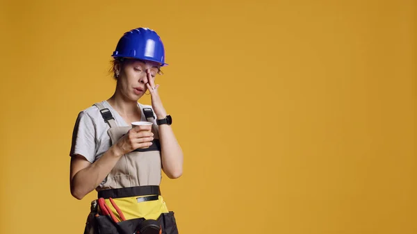 Portrait of sleepy handywoman drinking coffee cup at building work, feeling exhausted and falling asleep. Construction worker serving espresso refreshment and beverage over yellow background.