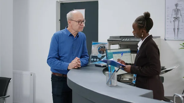 stock image Patient talking to female receptionist before attending appointment with doctor at medical facility center. Woman helping old man with checkup report papers at hospital reception desk.