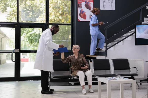 診察中にサインをするために高齢者に医療文書を与える医師は 病院の待合室で予約を訪問します 相談前に病気の症状を説明するシニア女性 — ストック写真
