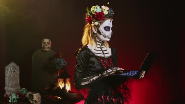 ハロウィンの衣装を着た不気味な女性がラップトップを持ち スタジオで伝統的なメキシコの休日にポーズをとっています 死者の日に頭蓋骨を身に着けていることは インターネットを閲覧するためにワイヤレスPcを使用して 体の芸術を構成する — ストック動画