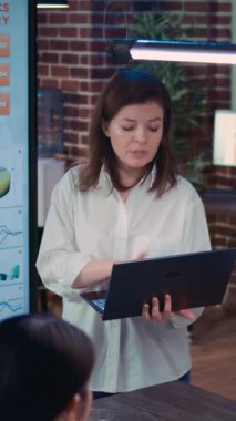 Dikey video: İş görüşmesi sunumunda konuşan çalışan, dizüstü bilgisayarı tutan, mali analiz raporunu tutan. Ekip şirket araştırma istatistikleri üzerinde çalışıyor, pazarlama stratejisi planlıyor, tartışıyorlar