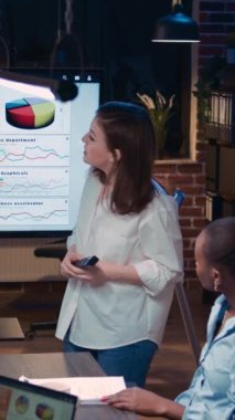 Dikey video: İş analizi ekip toplantısı, kurumsal sunum gösteren kadın, pazarlama stratejisi planlama. Satış istatistikleri dijital kurulda, iş arkadaşları ofiste beyin fırtınası yapıyor.