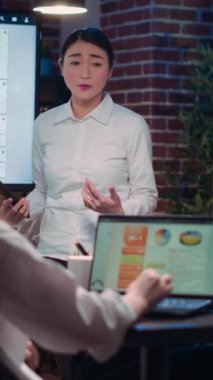 Dikey video: İş toplantısında analitik araştırma sunumu yapan Asyalı iş kadınları dijital ekranda istatistik şemalarını gösteriyorlar. Ofis çalışanı pazarlama raporu, strateji planı