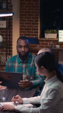Dikey video: İş arkadaşları beyin fırtınası yapıyor, Afro-Amerikan takım analisti dizüstü bilgisayar ekranında gelir istatistikleri gösteriyor. İş toplantısında strateji planlayan, analitik sunumunu tartışan insanlar