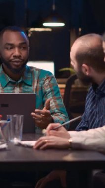 Dikey video: Afro-Amerikan analitik uzmanı iş toplantısında laptop ekranında veri görselleştirmesi gösteriyor. Takım çalışması, çeşitli iş arkadaşları şirket raporlarını gece vakti tartışıyor.