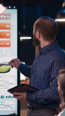 Dikey video: Şirket analisti istatistik şemalarını açıklıyor, iş toplantısında sunum yapıyor, dijital tablet tutuyor. Geceleri ekranda analitik araştırma çizelgeleri gösteren bir çalışan