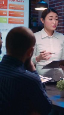 Dikey video: Asyalı şirket analisti iş toplantısında konuşuyor, finansal rapor sunumu, elinde pano tutan kadın. Çeşitli takımlar gelir istatistiklerini tartışıyor, konuşuyor, satış stratejisi planlıyor