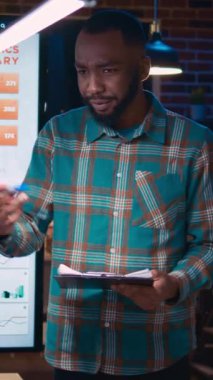 Dikey video: Afrikalı Amerikalı yönetici şirket finans raporunu sunuyor, çalışan çalışma alanında konuşuyor. İş toplantısındaki çeşitli iş arkadaşları, gelir istatistikleri sunumu