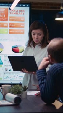 Dikey video: Çalışan iş toplantısında konuşuyor, finansal rapor sunumu, analitik grafiklerle dijital panonun yanında duruyor. Şirket araştırma istatistikleri üzerinde çalışan ekip, pazarlama planlaması