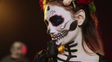 Stüdyonun arka planında ölüm kostümü ve çiçek çelengi giyen göz kamaştırıcı tanrıça, kutsal noel baba gibi davranıyor. La Cavalera Catrina Kafatası vücut sanatıyla ölü kadın, Meksika kültürü. El kamerasıyla..