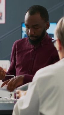 Dikey video: Afrikalı Amerikalı bir adam son sınıf öğrencisiyle ilaç ürünleri hakkında konuşuyor, eczanedeki ilaçlar ve haplar hakkında sorular soruyor. Eczanedeki eczacılara bakıyorum..